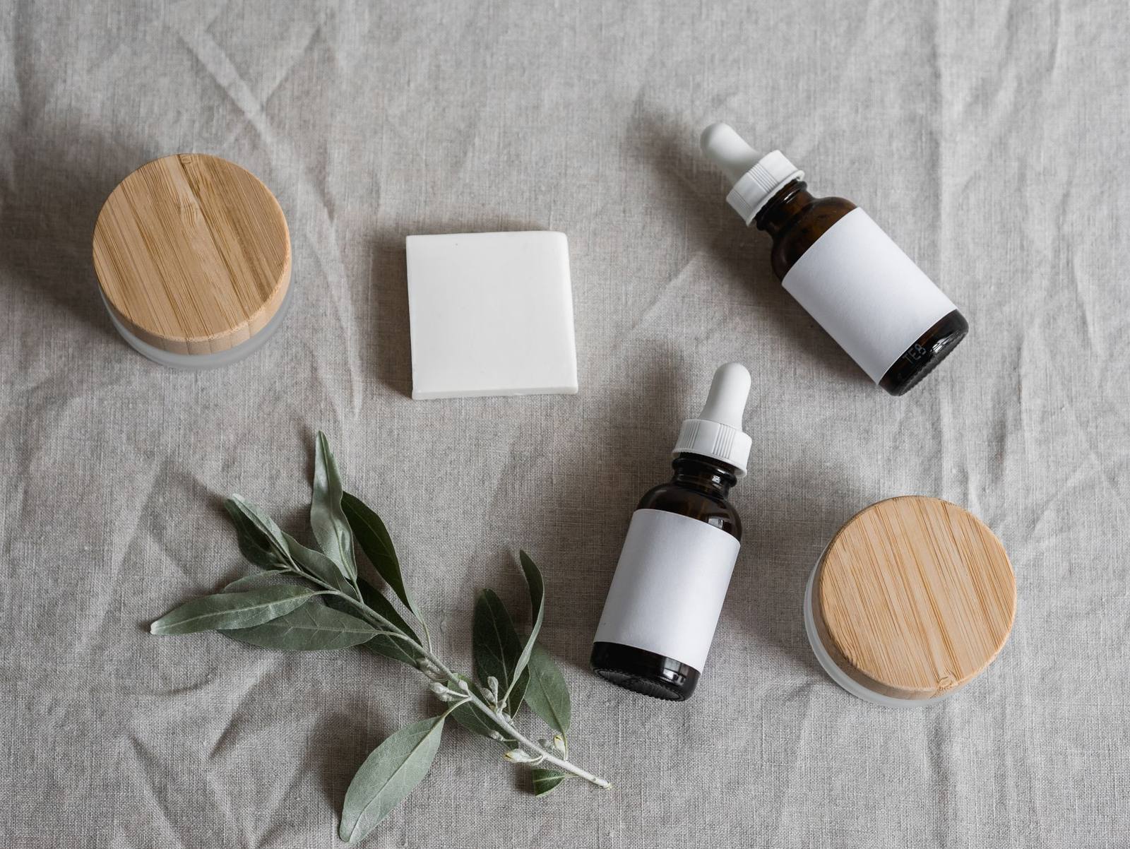 Plastic free skincare packaging: glass dropper bottles with wooden lids and olive leaves on linen